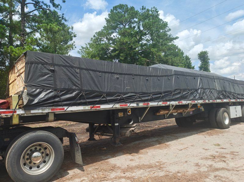 Flatbed trailer carrying a long black tarp-covered load outdoors under a cloudy sky