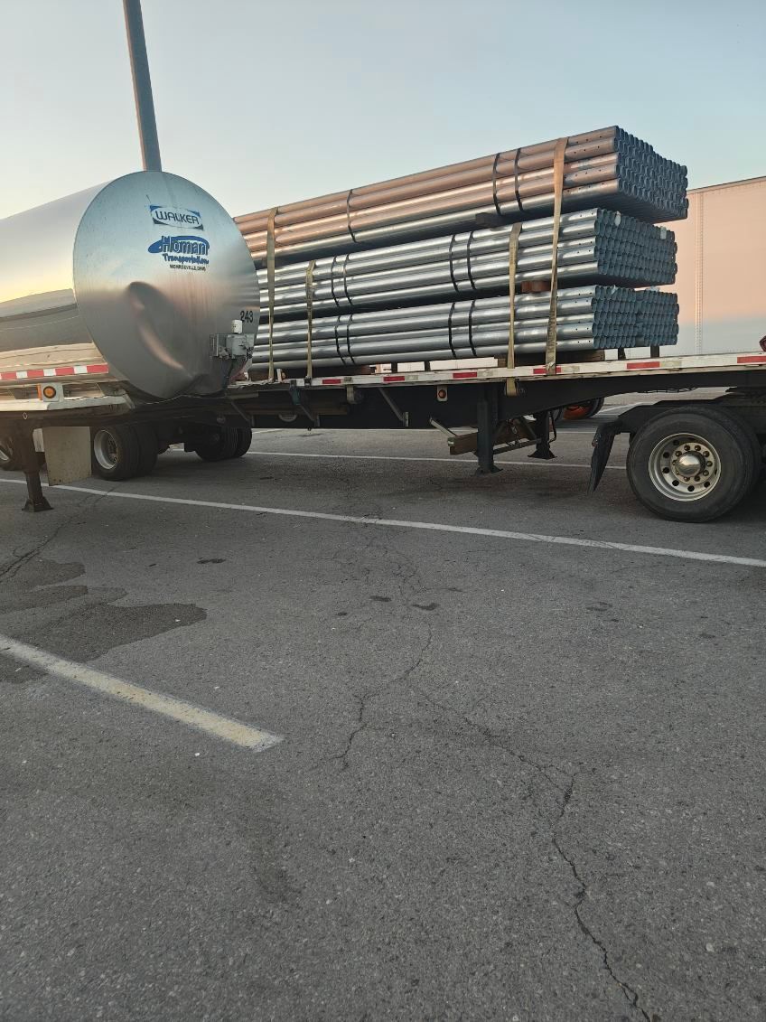 Loaded tanker truck beside a flatbed trailer carrying stacked metal pipes in a parking lot