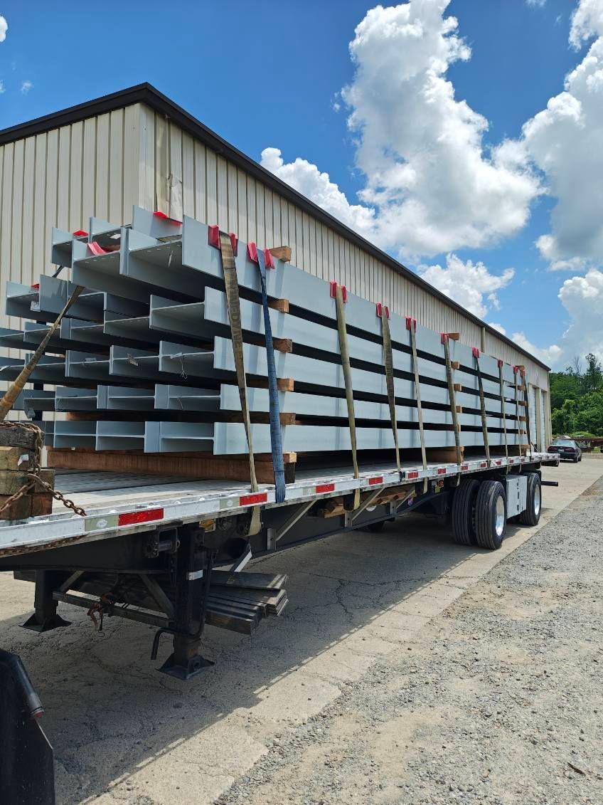 Flatbed trailer loaded with stacked metal beams beside a warehouse on a gravel lot