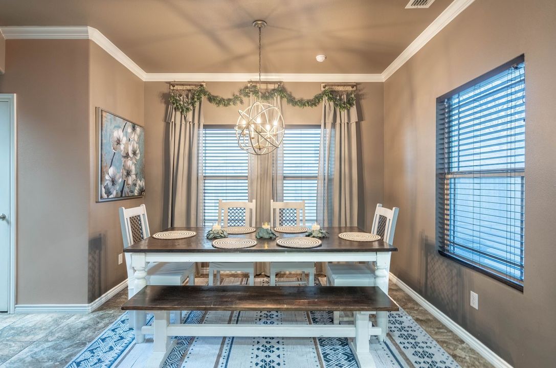 Dining room with a long wooden table, blue rug, hanging greenery, and bright windows with blinds.