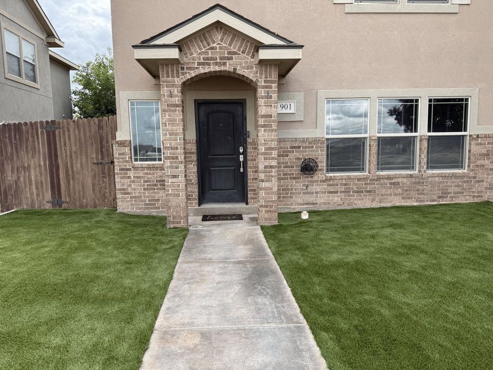 Front view of a beige house with a black front door, brick entryway, windows, and a concrete walkway across the lawn