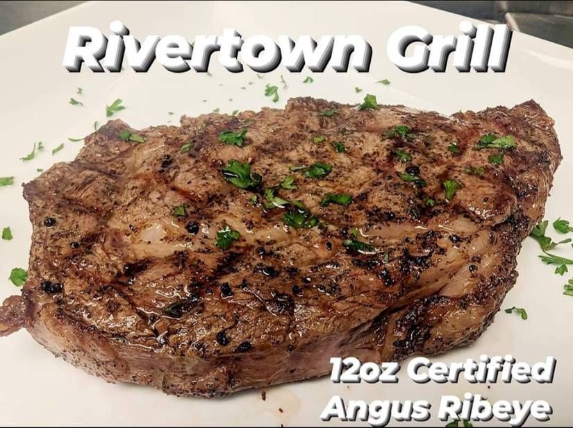 Grilled steak on a white plate with parsley, labeled “Rivertown Grill” and “12oz Certified Angus Ribeye”