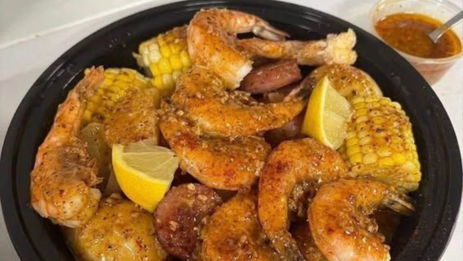 Seasoned shrimp, corn on the cob, and sausage on a black platter with lemon wedges and dipping sauce