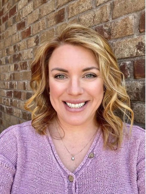 Smiling person in a lavender sweater standing before a brick wall