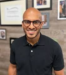 Smiling man in black polo and glasses standing indoors with framed photos on the wall