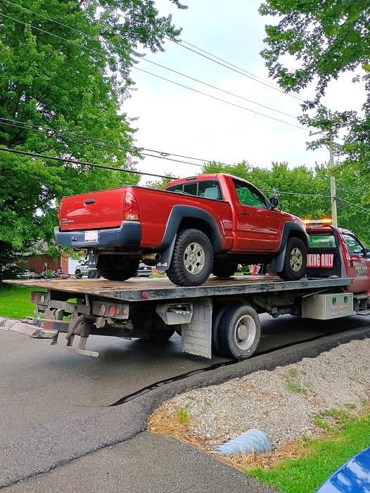 Red pickup truck strapped on a tow truck on a tree-lined street.