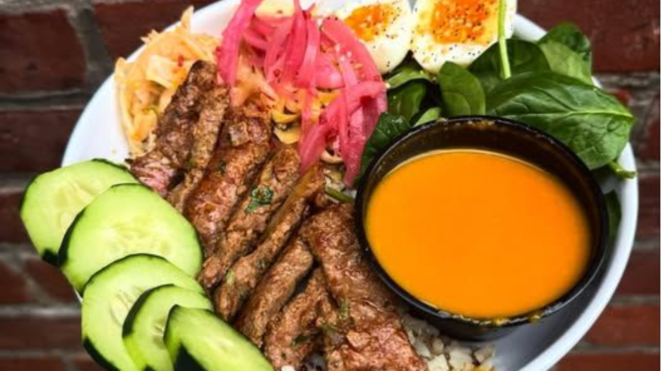 Plated grilled meat with avocado, pickled vegetables, eggs, greens, and orange dipping sauce on a table