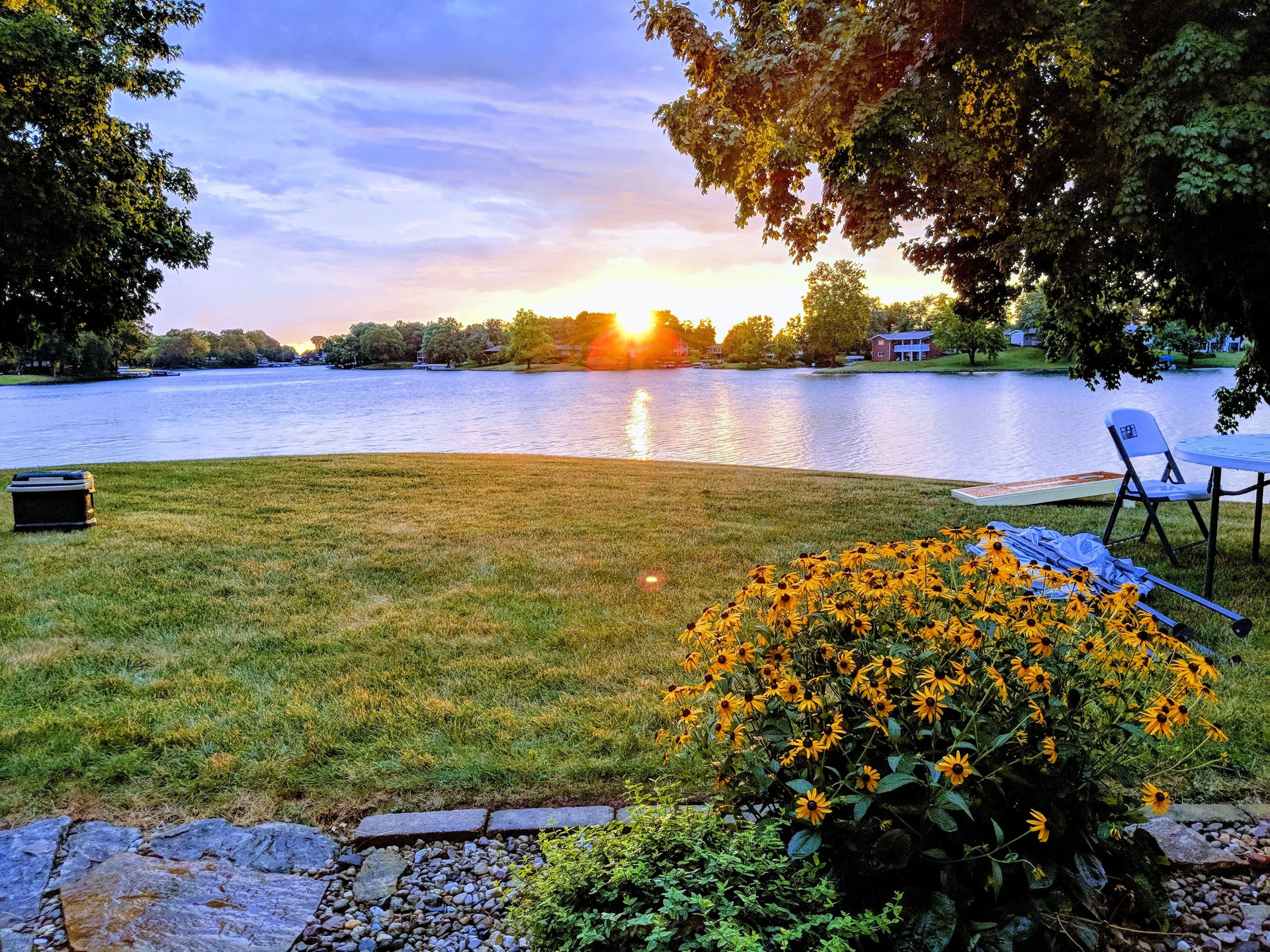 Sunset over a lake; golden light reflects on the water. Yellow flowers and green grass in the foreground.
