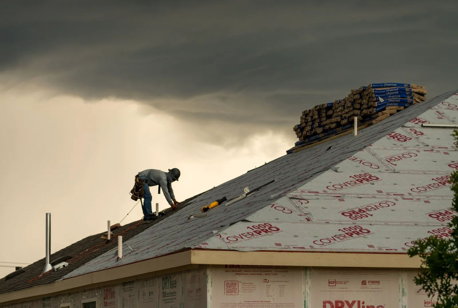 A worker installs new shingles on a steep roof beneath dark storm clouds during a roof replacement. A worker installs new shingles on a steep roof beneath dark storm clouds during a roof replacement.