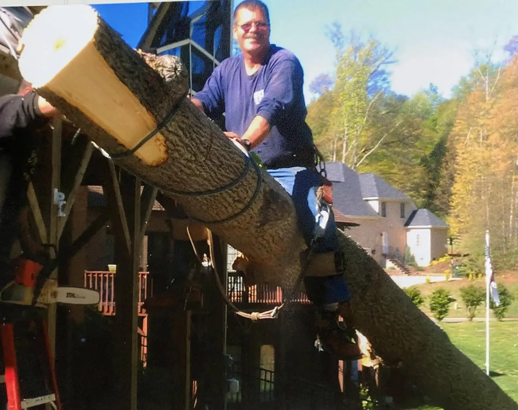 Man sits on a log, attached by a rope, using a chainsaw. Building and trees in the background.