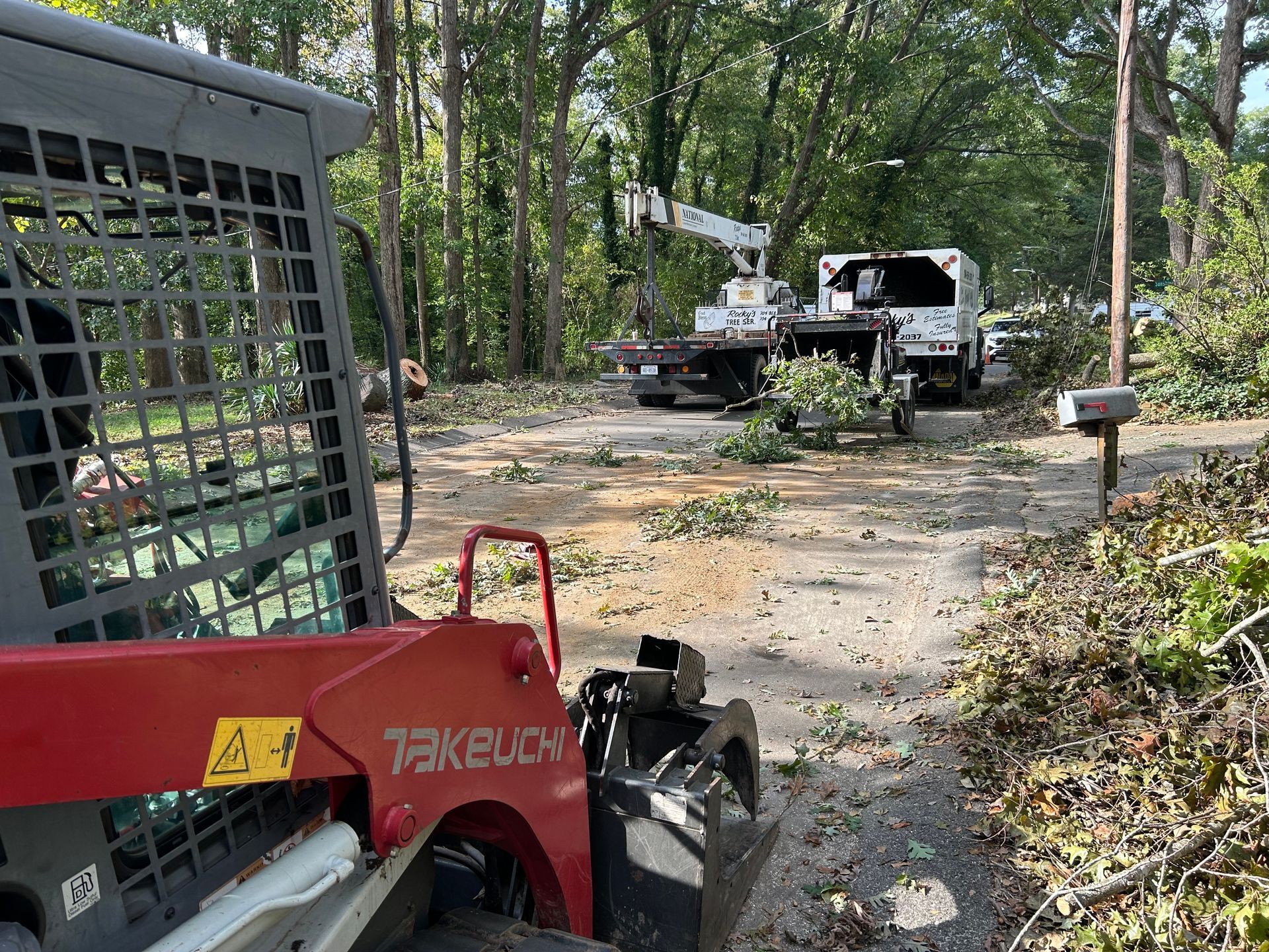 Skid steer loader and utility trucks clearing debris from a driveway with trees in the background.