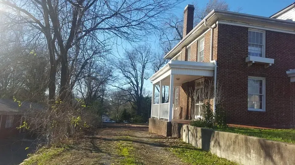 Two-story brick house with a porch. A driveway leads to the house, trees line the left. Sunny day.