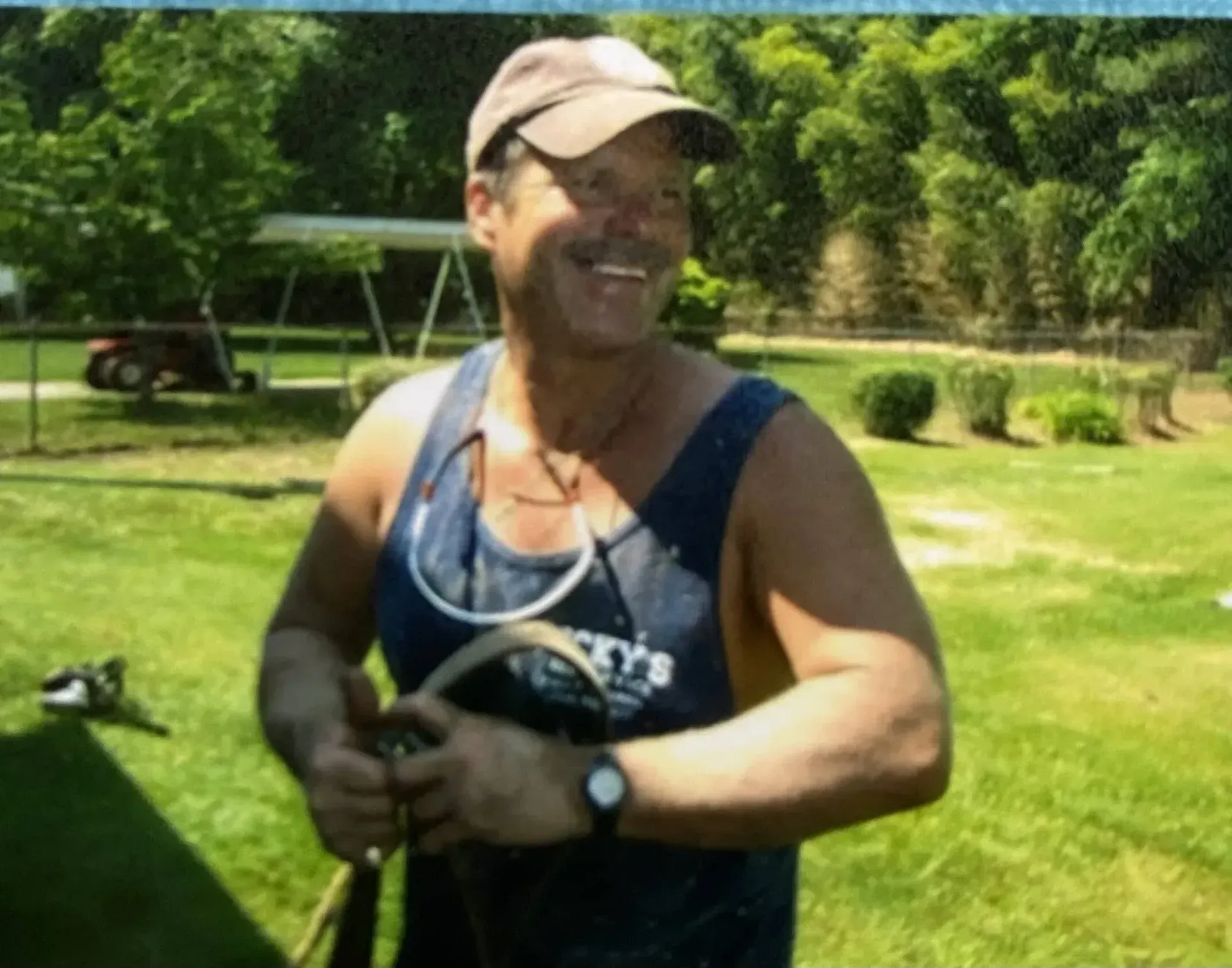 Man in a tank top smiles outside; holds tool, wears hat, watch. Green lawn, trees in background.
