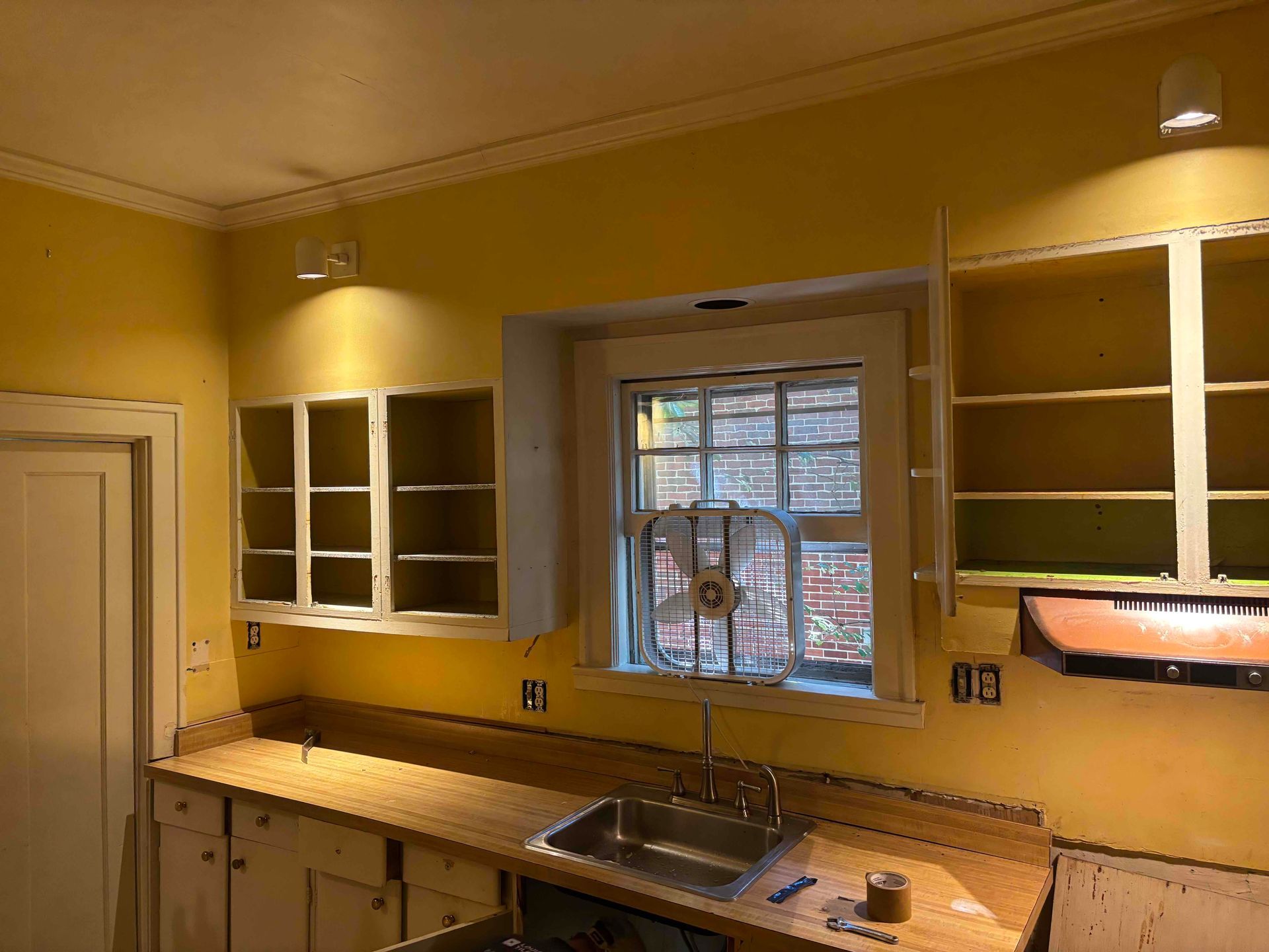 A kitchen renovation in progress, featuring yellow walls, open cabinets, a sink, and a window fan.