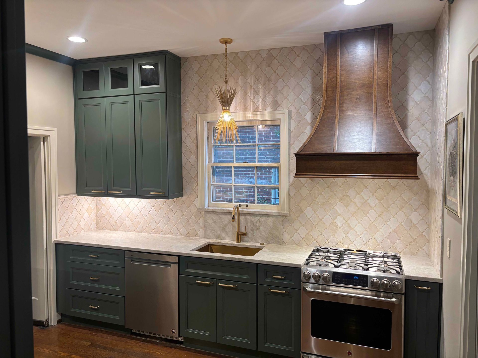 A kitchen with dark green cabinets, white countertops, a large wooden range hood, and a decorative tile backsplash.
