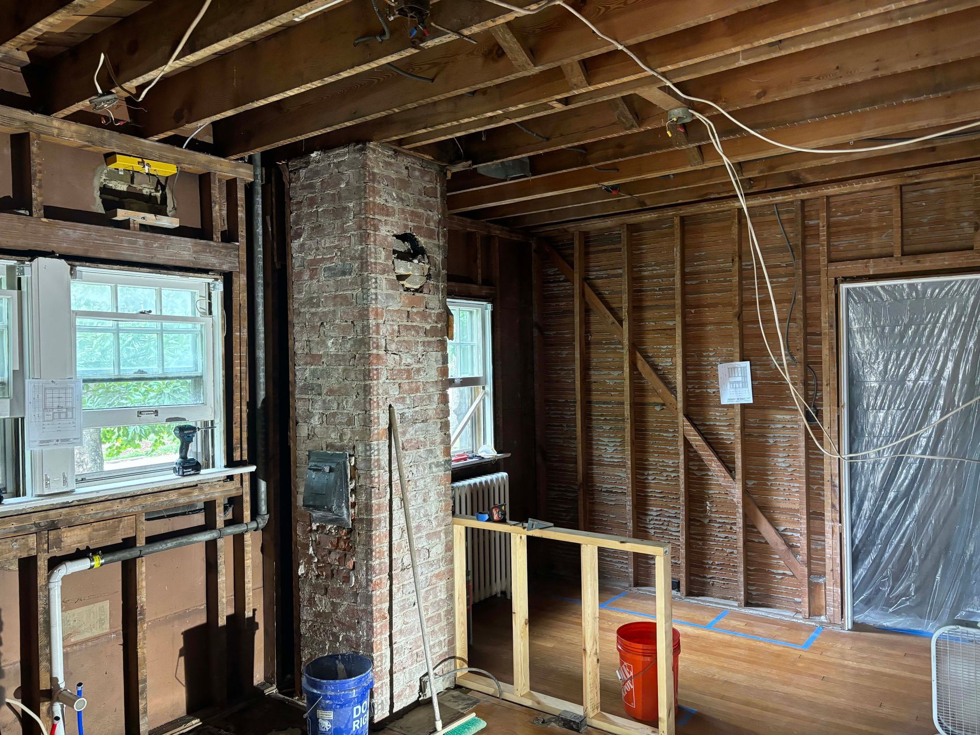 Interior view of a room under renovation with exposed wood studs, an exposed brick chimney, and open ceiling joists.