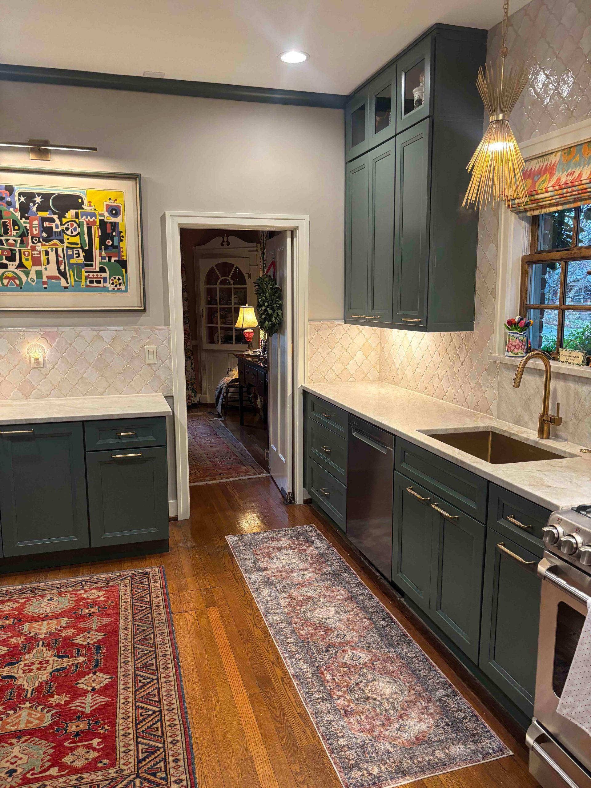 A kitchen with dark green cabinets, light stone countertops, a patterned runner rug, and a colorful abstract painting.