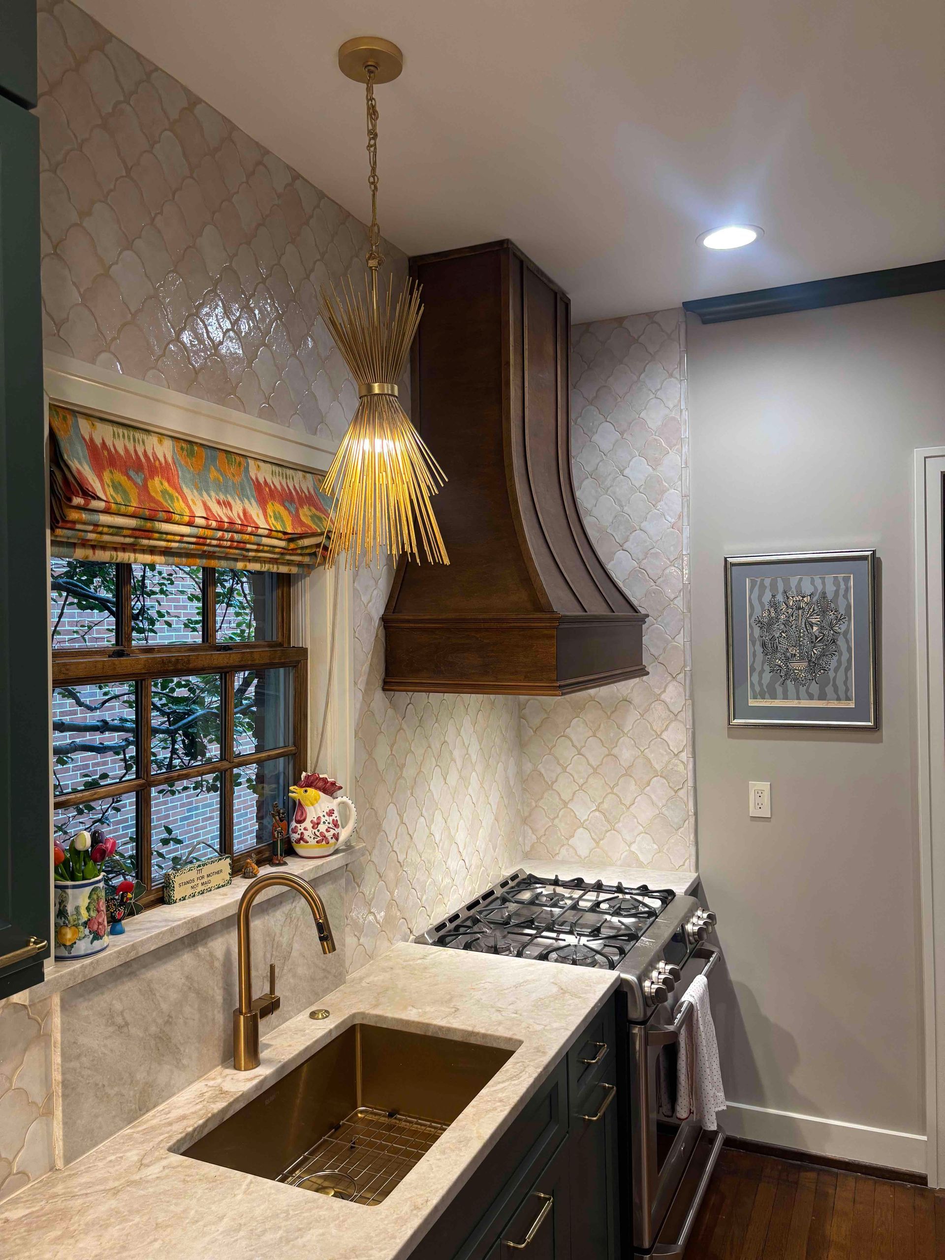 A kitchen corner with a wooden hood over a stove, a brass sink, patterned window shade, and an ornate gold pendant light.
