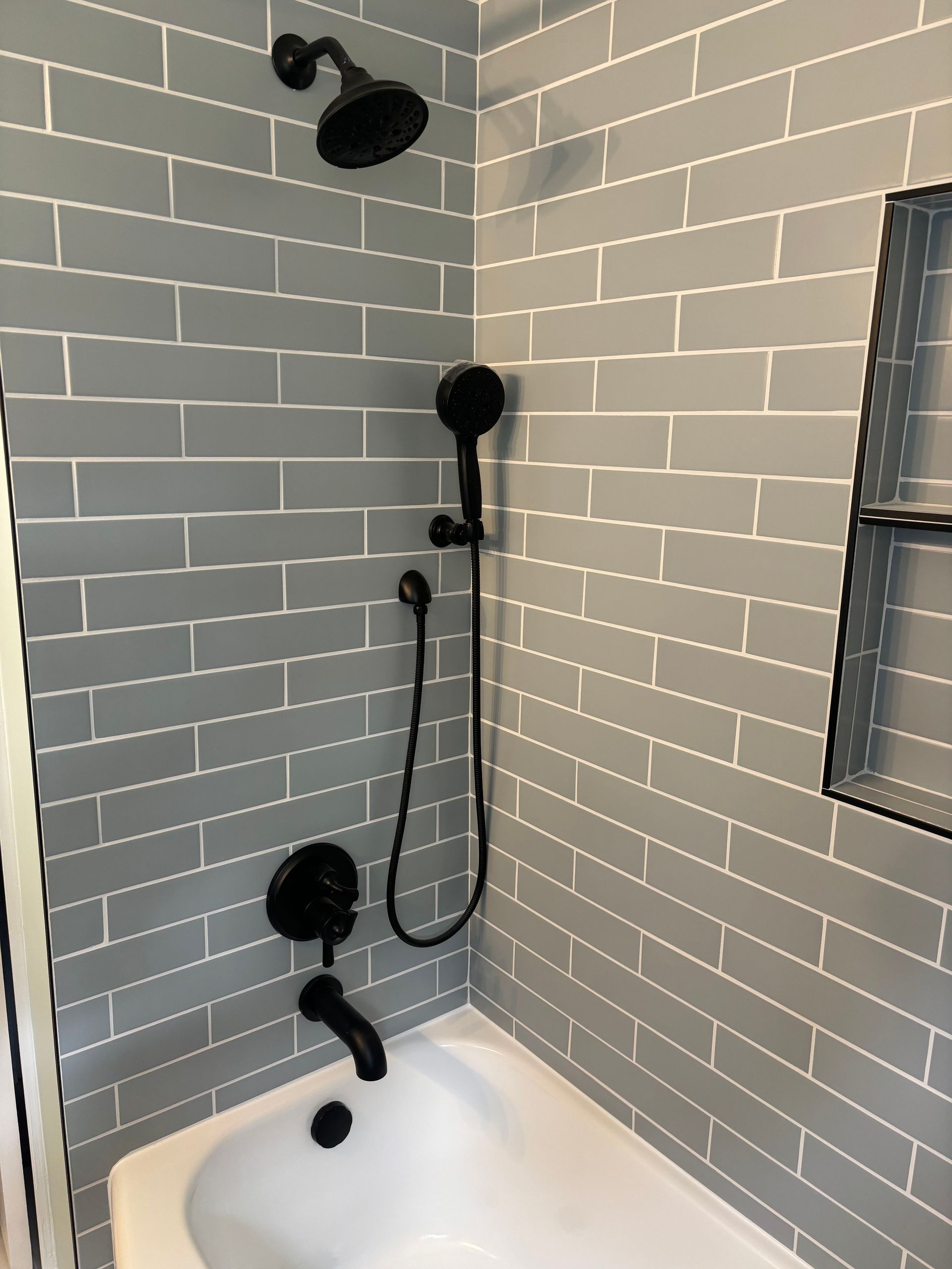 A bathroom shower area with grey subway tiles, matte black fixtures, a handheld sprayer, and a built-in recessed shelf.