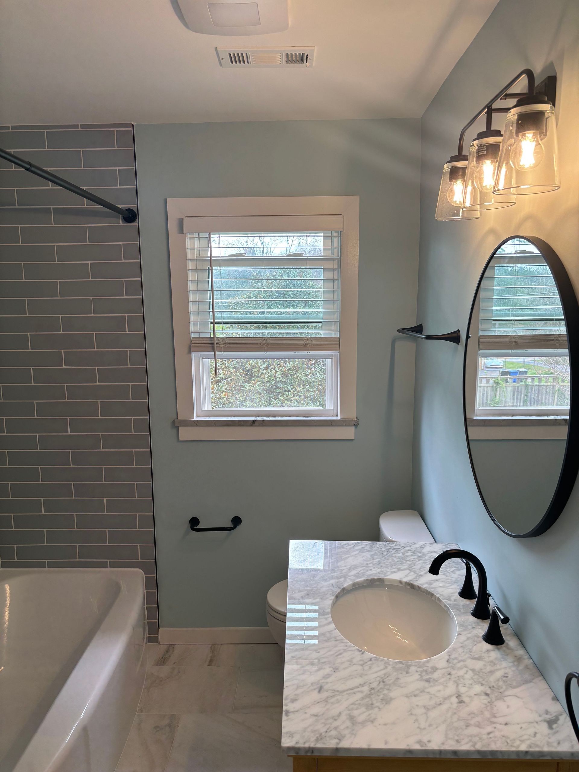 A bathroom with a gray tiled shower, light blue walls, a marble vanity, a round mirror, and a window with white blinds.
