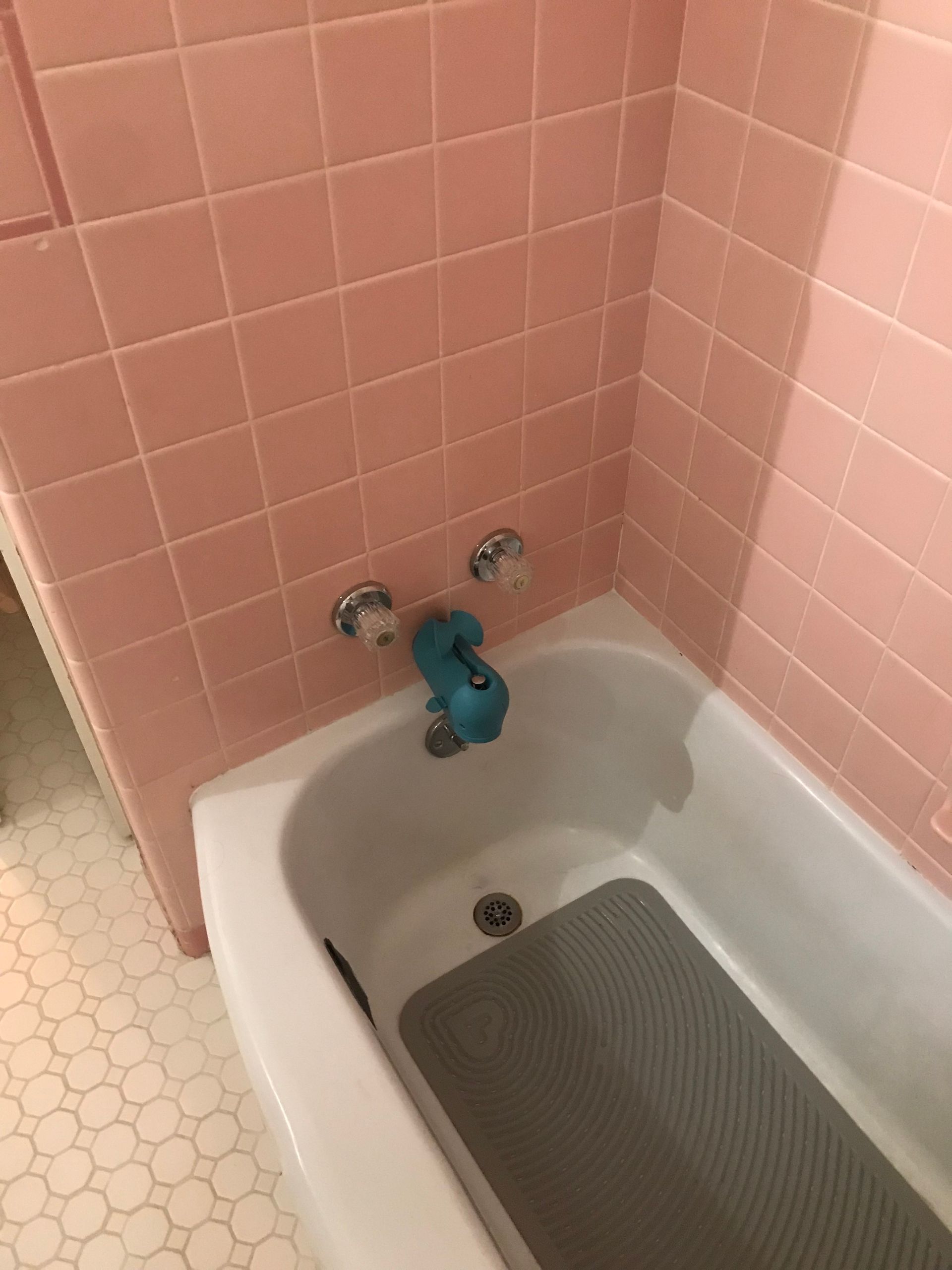 A white bathtub with a blue spout and two silver faucets, set against pink tiled walls with a gray non-slip bath mat inside.