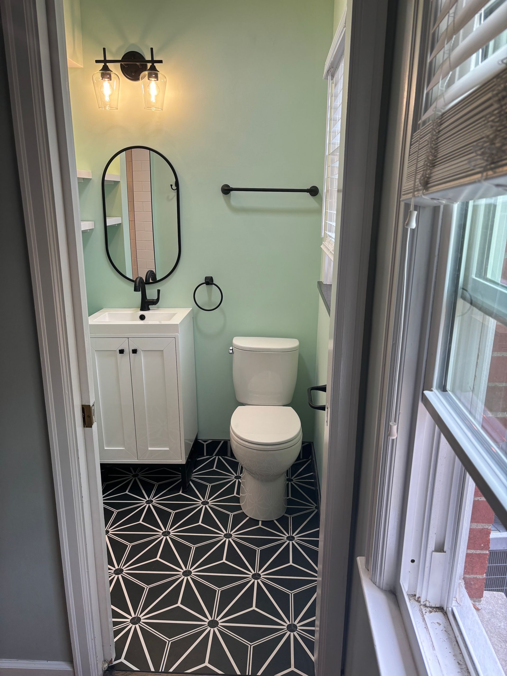 Bathroom with black and white patterned floor, white vanity, oval mirror, and light green walls.