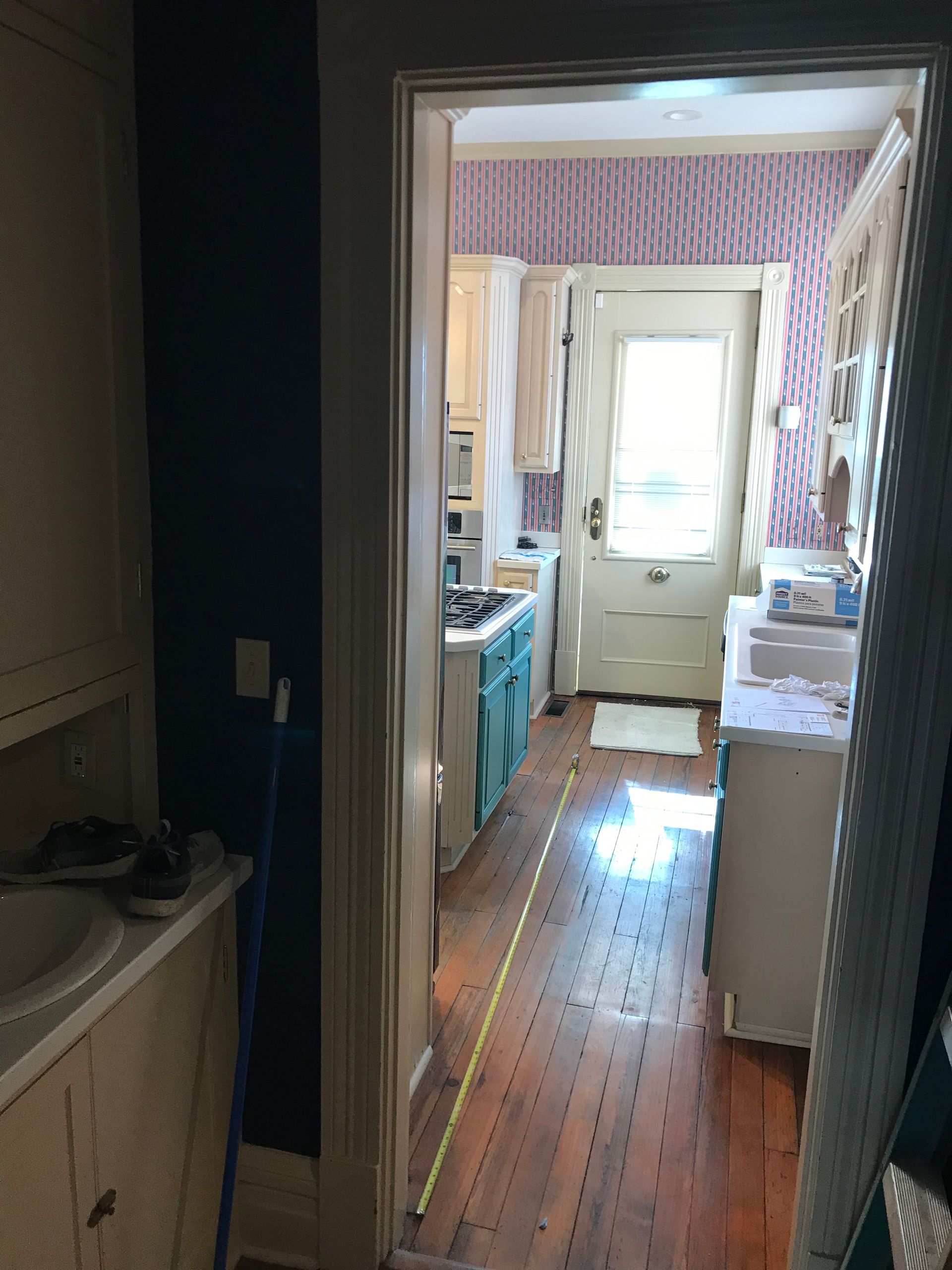 A view through a doorway into a kitchen with hardwood floors, white cabinetry, a patterned wall, and a yellow tape measure.
