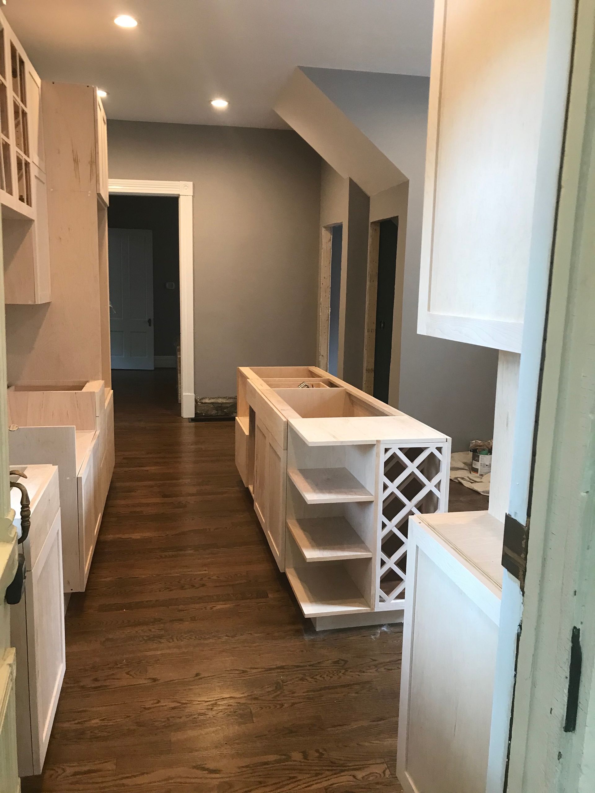 A kitchen under renovation showing wooden cabinets, a central island with wine storage, and hardwood floors.