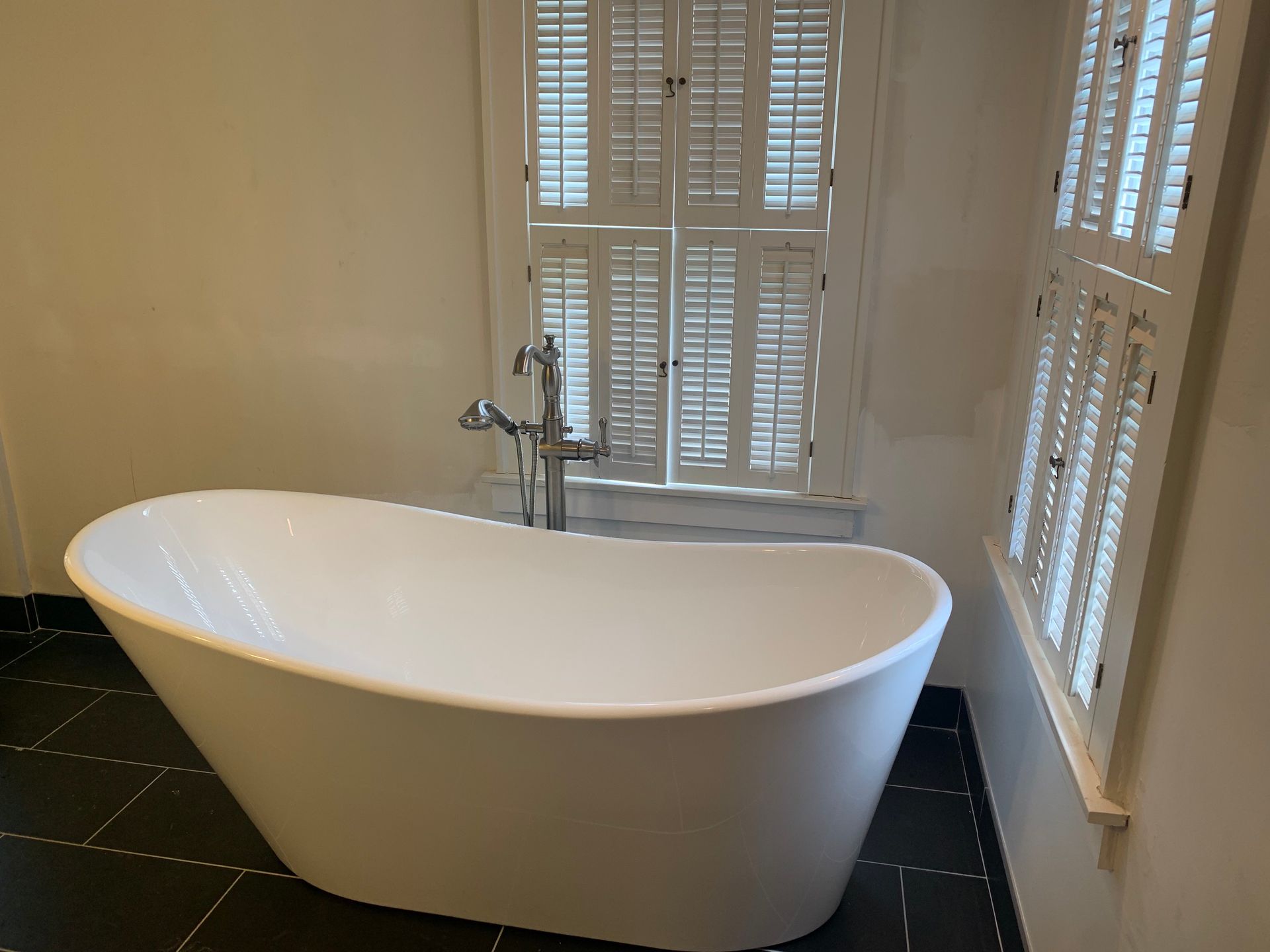 White freestanding bathtub in bathroom with shutters and dark tile floor.