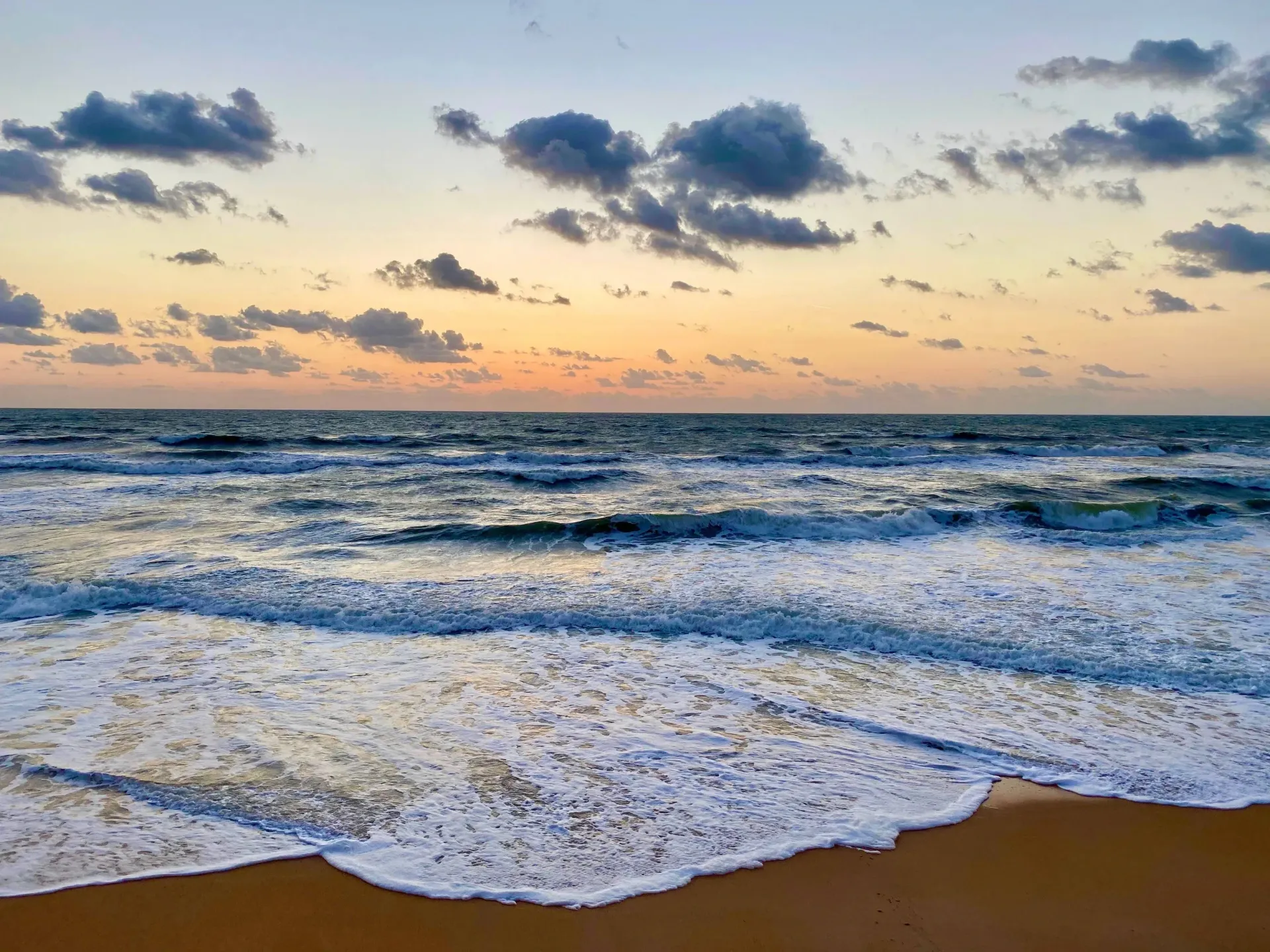 Sun over the ocean with waves on a sandy beach under a partly cloudy sky.