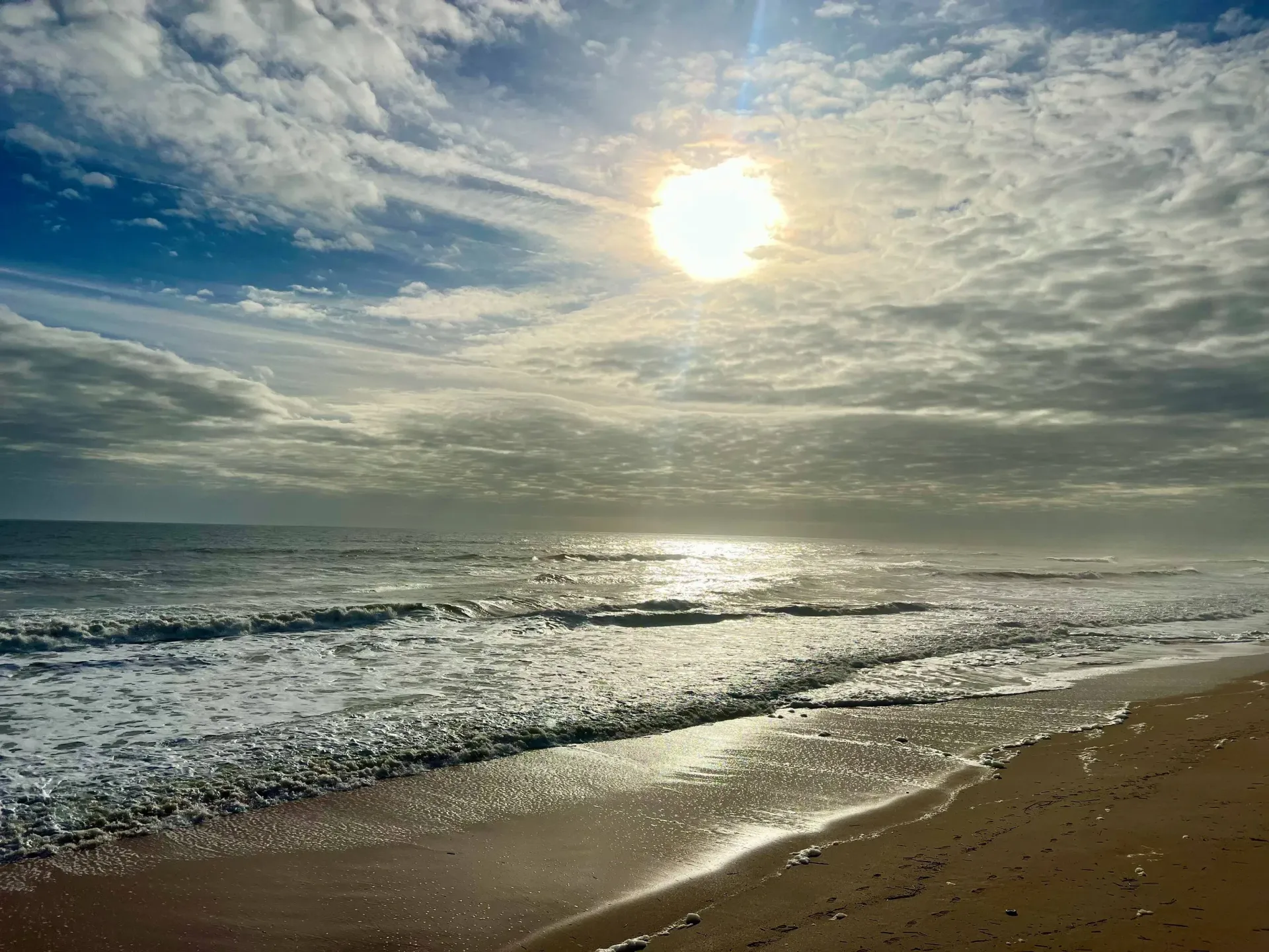 Partially cloudy sky with dazzling sun peeking through above the afternoon waves at the beach.