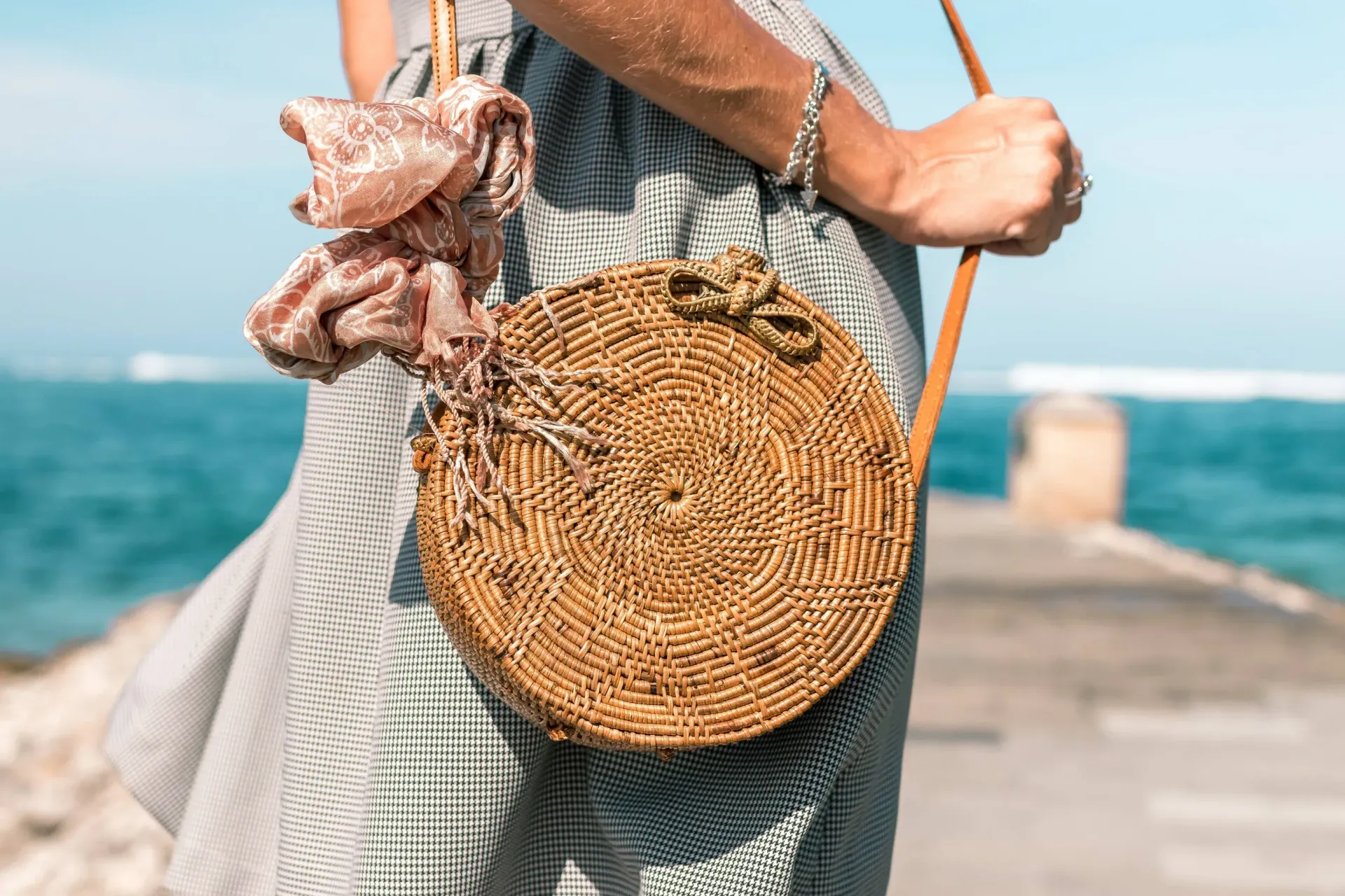 Person holding a round woven handbag with a pink scarf by the sea.