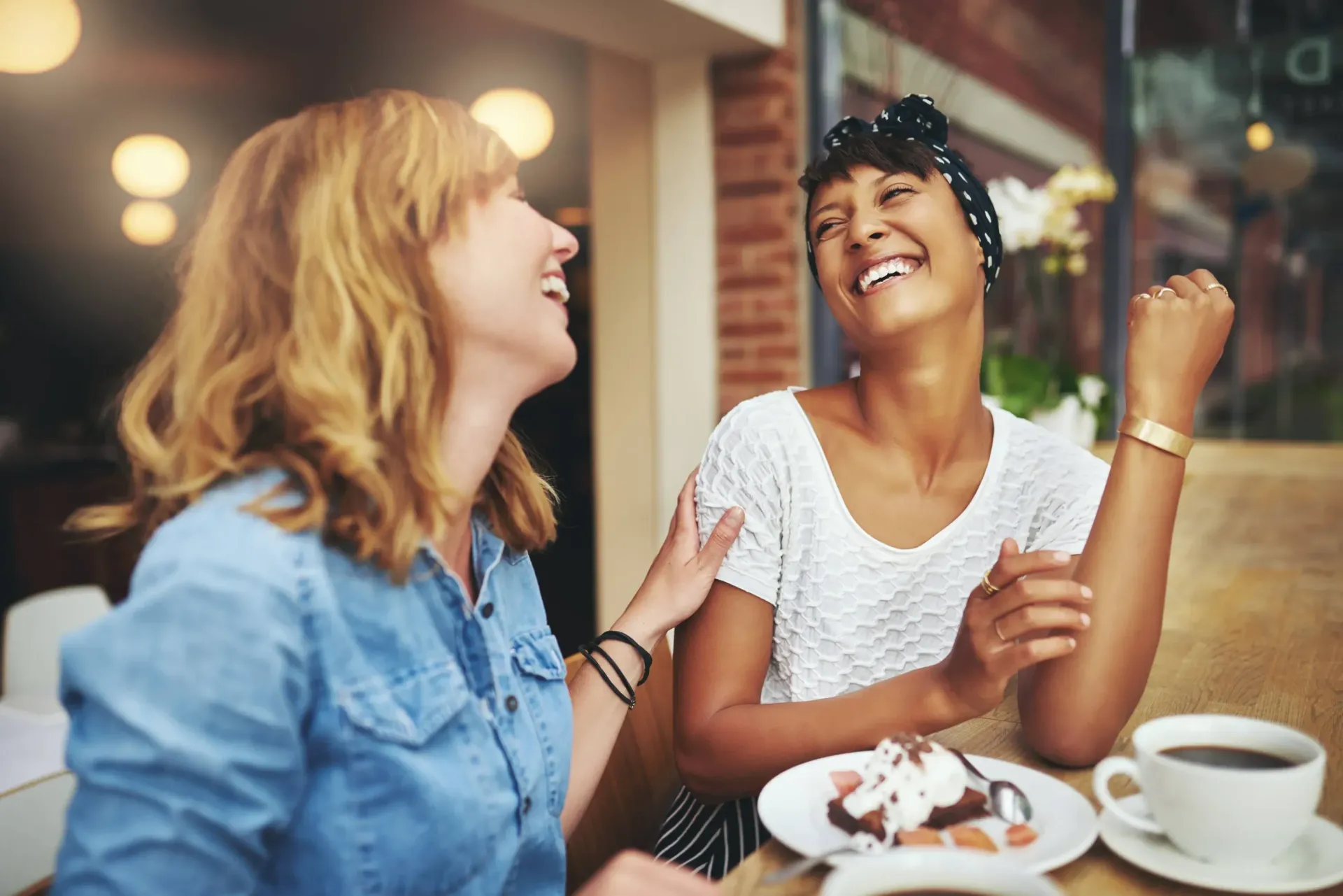 Lady friends laughing and enjoying dessert at a restaurant with outdoor seating.