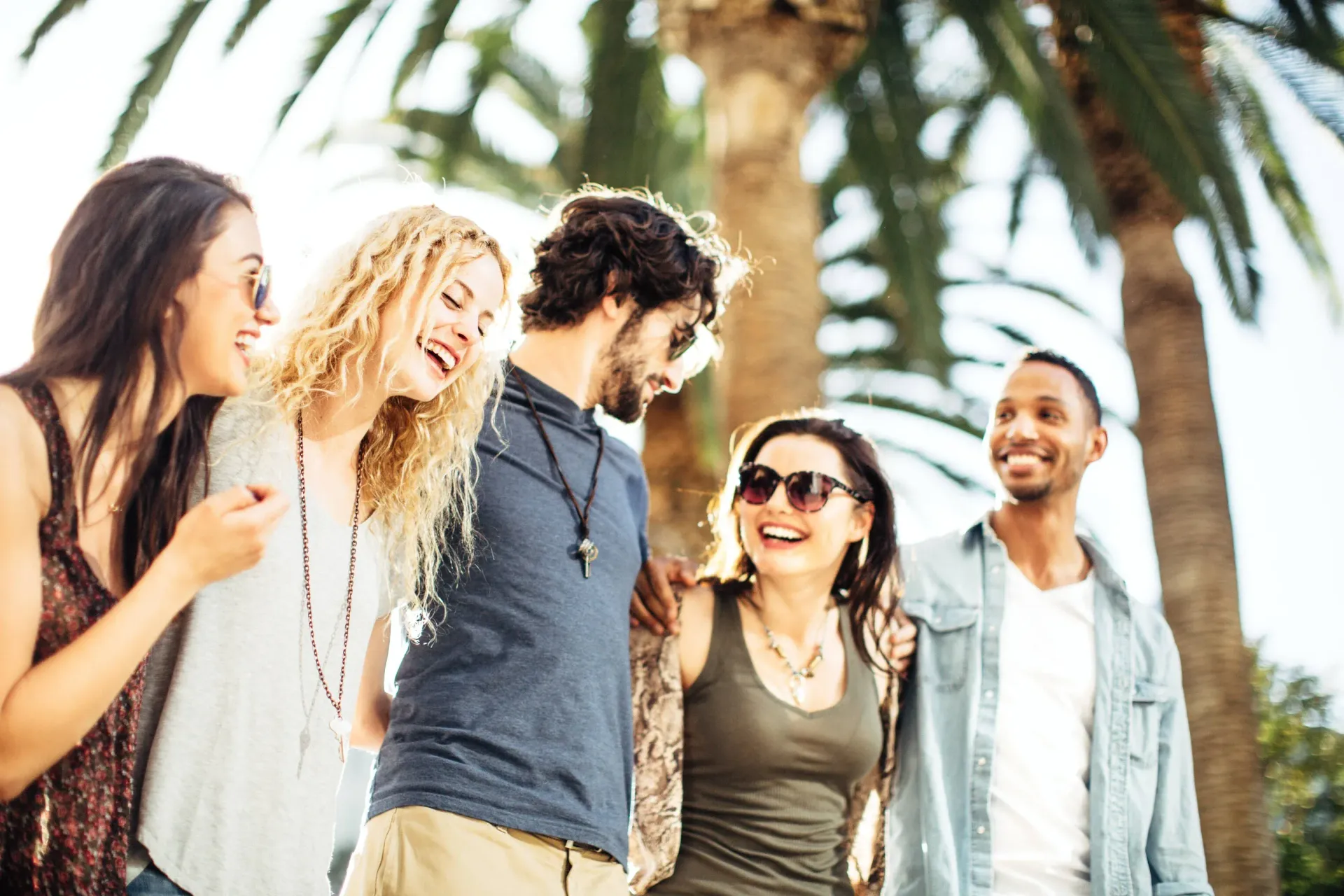 Group of friends laughing, arms around each other, outdoors near palm trees. Sunny day.
