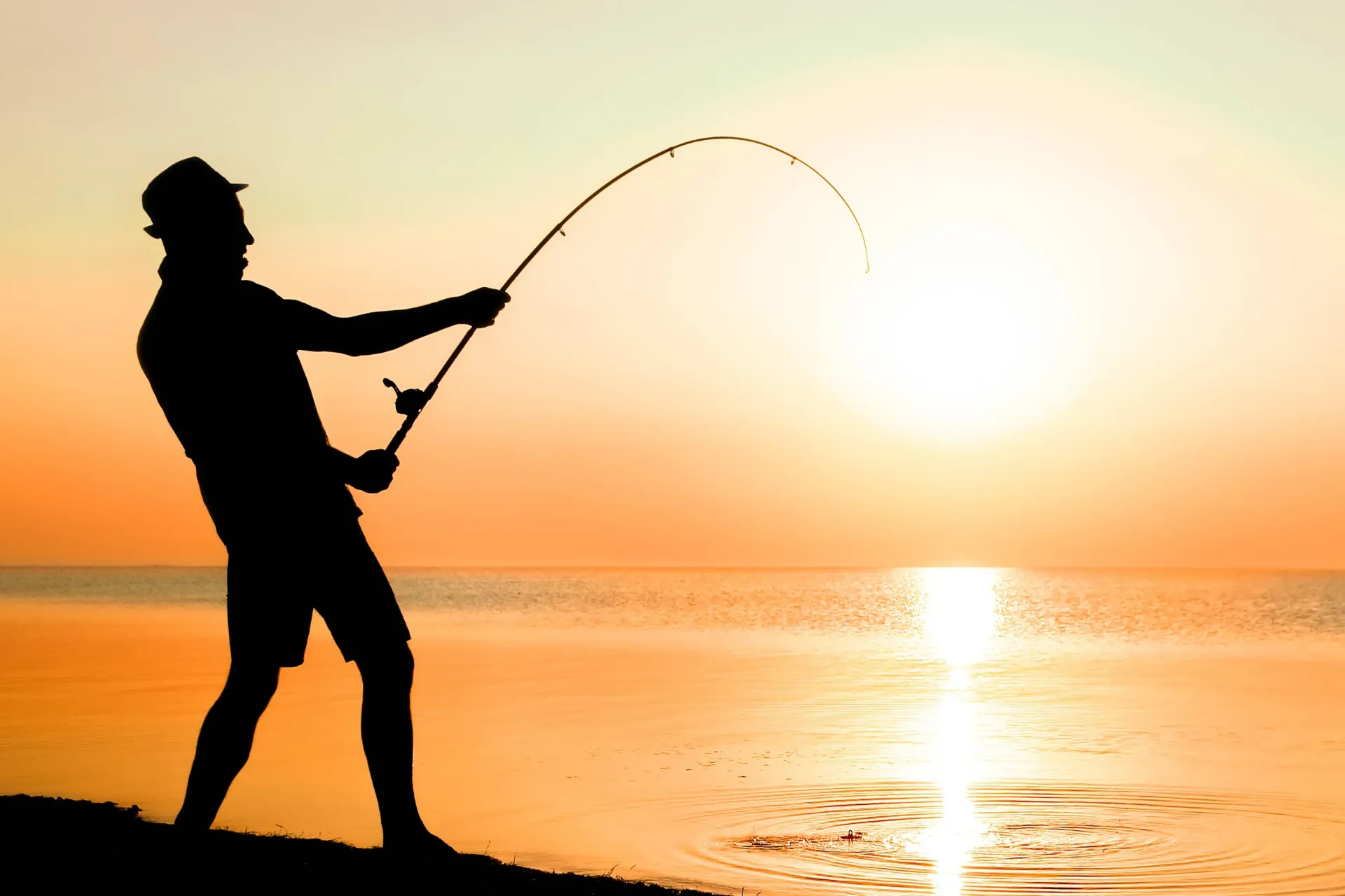 Silhouette of a person fishing at sunset by the water, with a glowing orange sky.