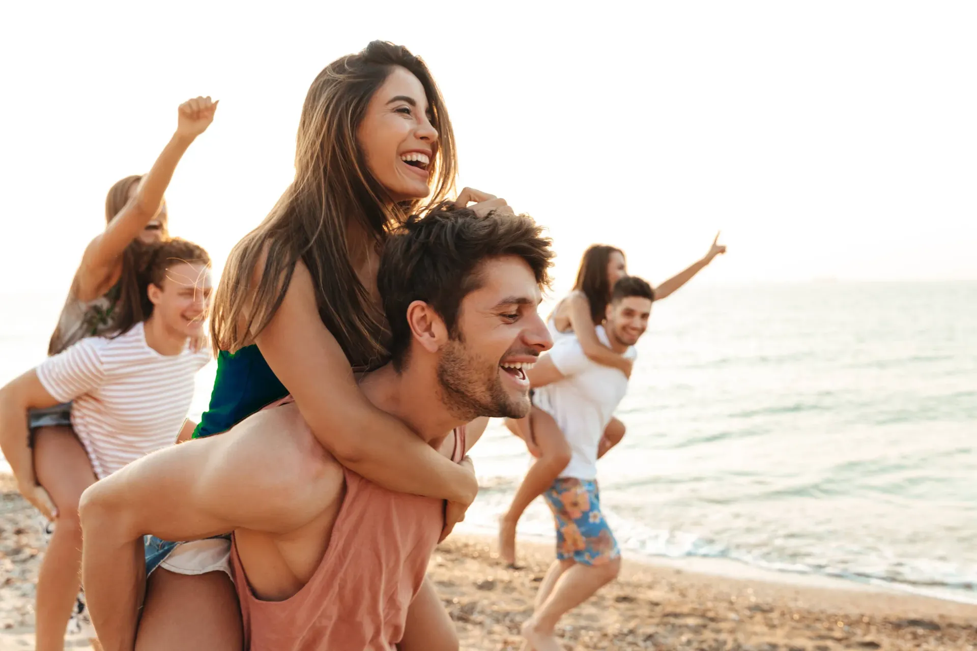 People having fun on a beach: two men carrying women on their backs, others cheering, all smiling in the sunlight.