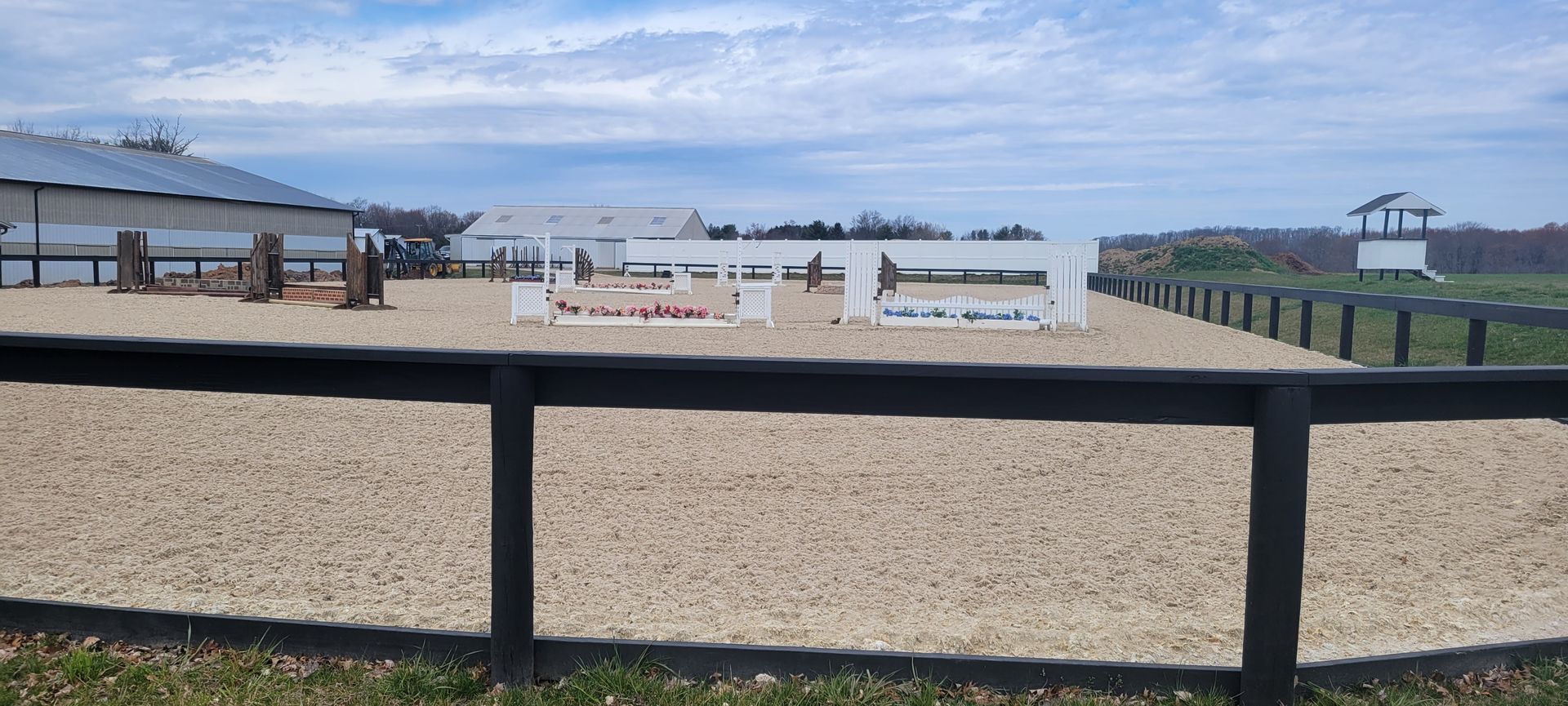A gravel horse riding arena with jumps, enclosed by a black wooden fence under a cloudy sky.