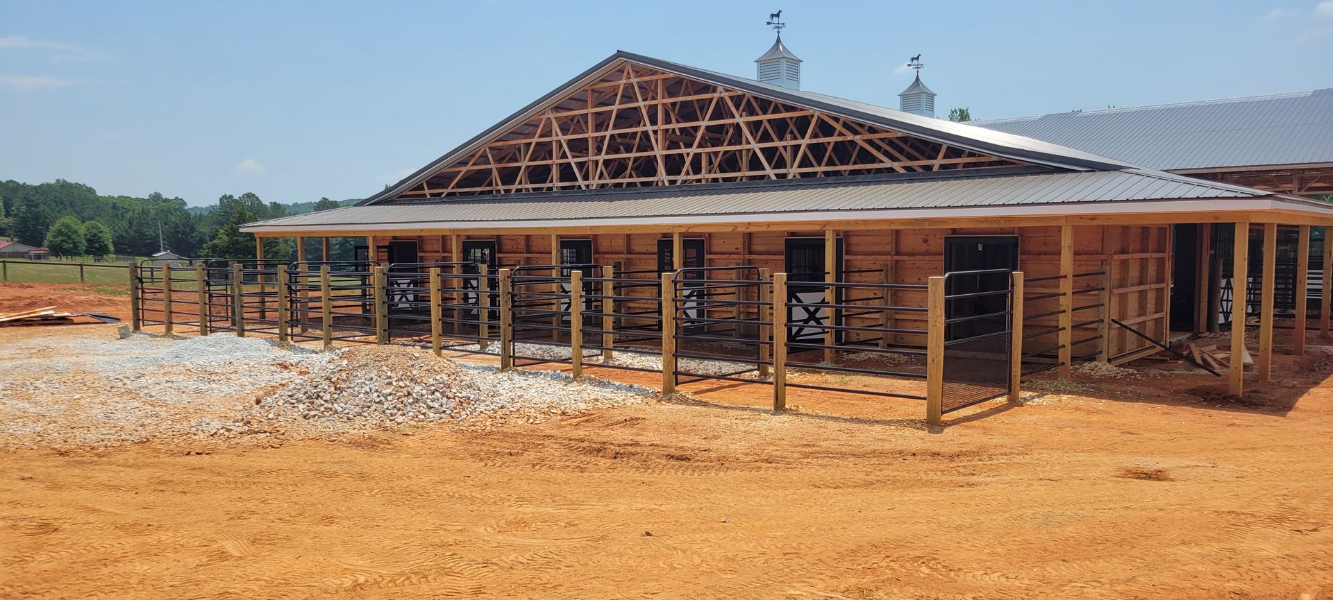 A partially constructed barn with a wood frame, metal roof, and surrounding dirt lot under a clear blue sky.