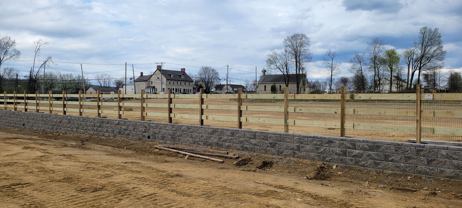 A low stone wall borders a dirt field in front of wooden fence posts, trees, and buildings under a cloudy sky.