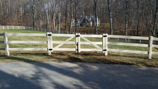 A white, three-rail wooden fence with a matching double gate in a grassy field with trees in the background.