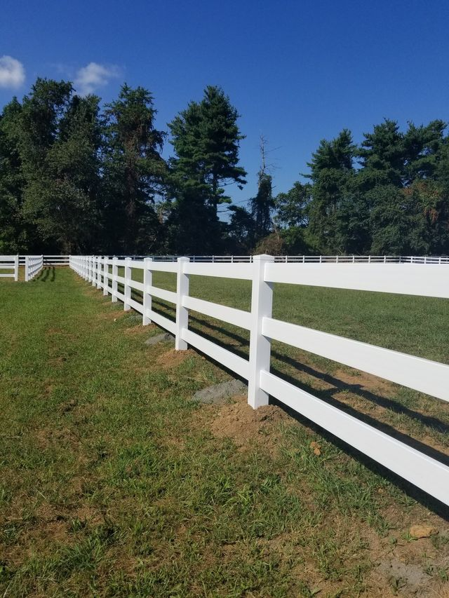 A long, white three-rail fence borders a green grassy field under a clear blue sky.