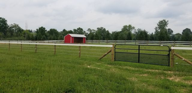 A grassy field with a metal gate in the foreground, white fencing, and a small red shed in the distance.