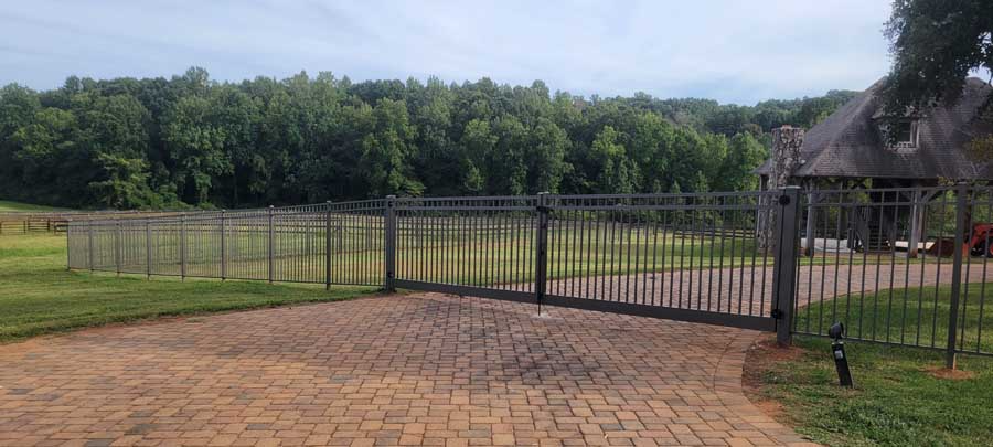 A brick driveway leading to a metal gate and fence in front of a grassy field bordered by trees under a cloudy sky.