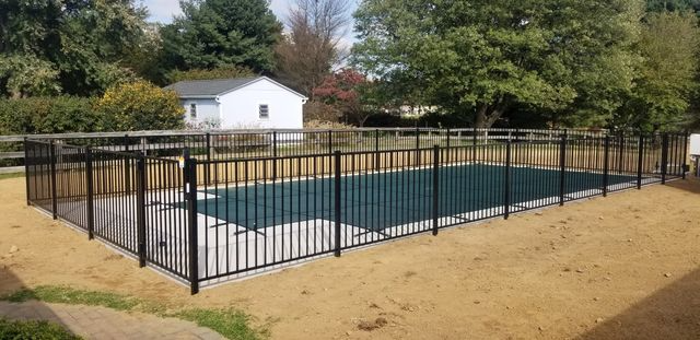 A black metal safety fence encloses a rectangular swimming pool covered with a dark mesh cover in a sandy yard.