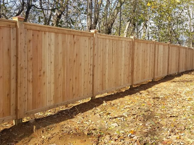 A long wooden privacy fence with vertical panels and square posts standing on a patch of earth covered in fallen leaves.