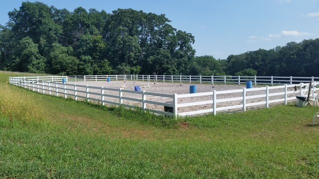 A fenced-in horse riding arena with sandy footing and blue barrels, set in a grassy field backed by trees.