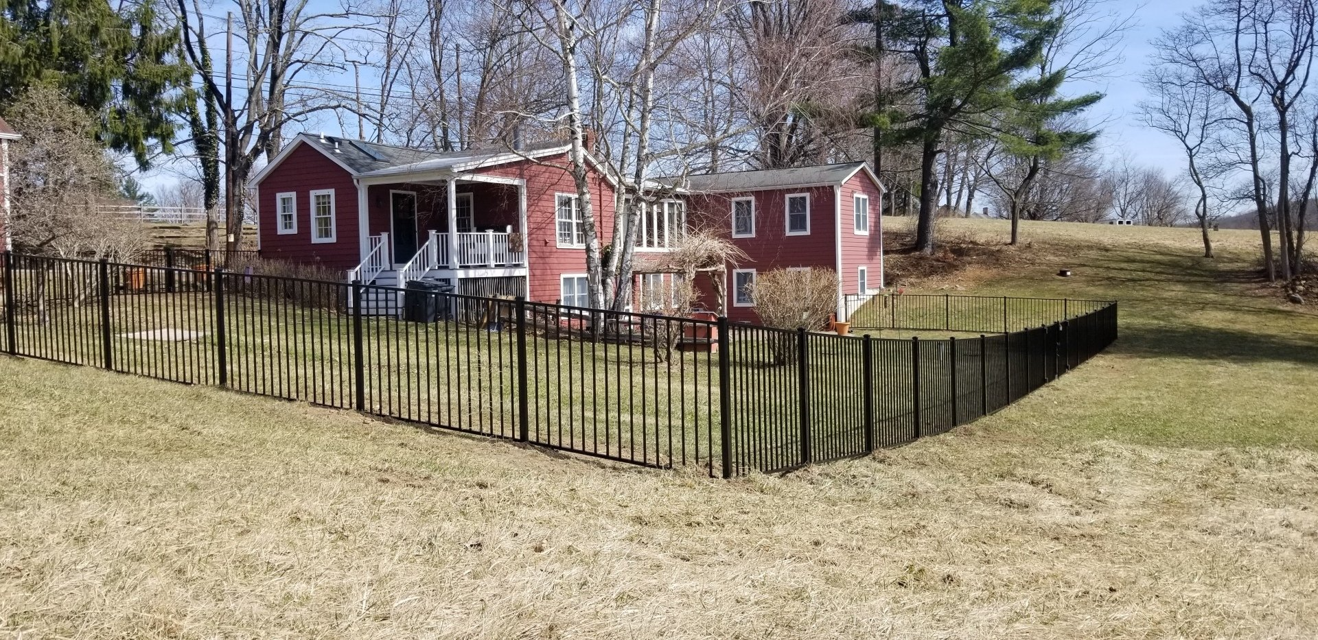 A red house with a white porch sits behind a tall black metal fence in a yard on a sunny day.