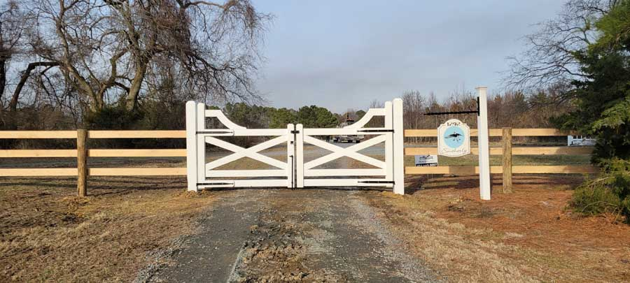 A white, double-swing gate with an X-pattern design, framed by a three-rail wooden fence on a rural dirt road.
