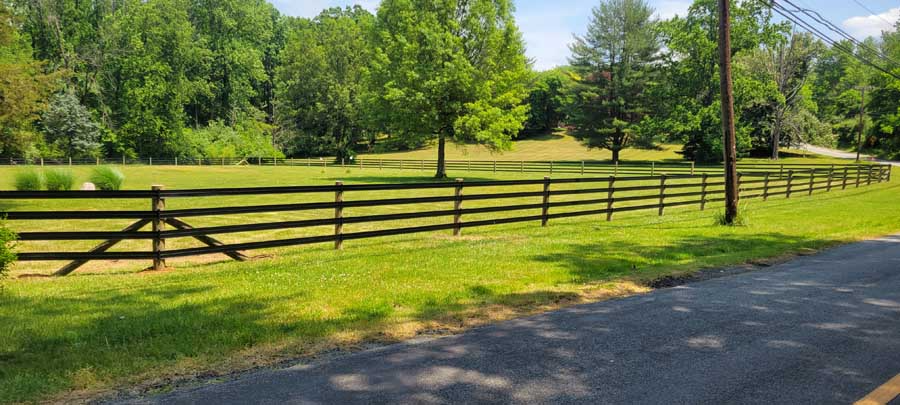 A wooden three-rail fence curves along a rural road, bordering a lush green field with scattered trees under a blue sky.
