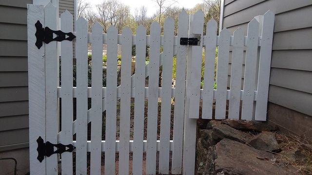 A white picket gate with black hinges hangs crookedly, leaving an open gap above a stone landscape border.